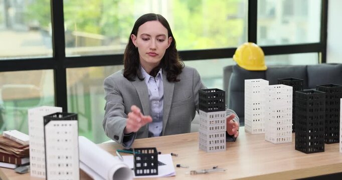 Lady architect assembles building models on desk checking facade layout. Designer aligns tower blocks adjusting structure height for project draft