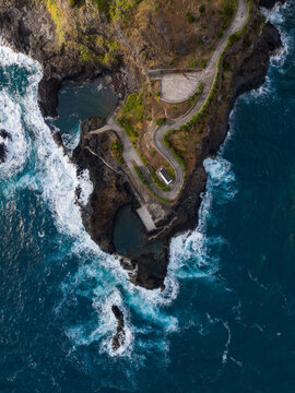 Aerial view of the rugged volcanic coastline featuring Seixal natural pools and a winding road along the dark cliffs with people on the shore in Seixal, Portugal.