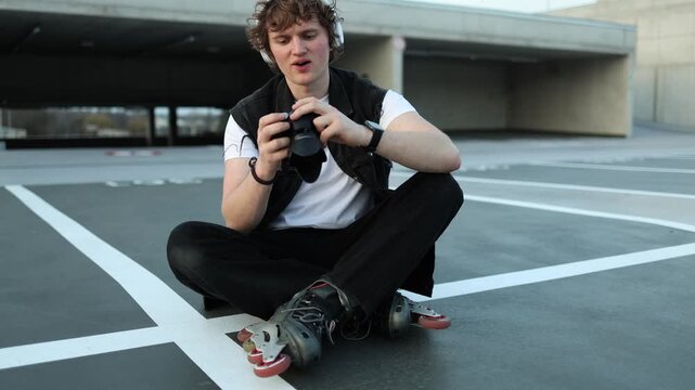 Young man on inline skates sits on empty rooftop parking and checks camera settings focused on hobby practice urban lifestyle creativity learning process and evening atmosphere