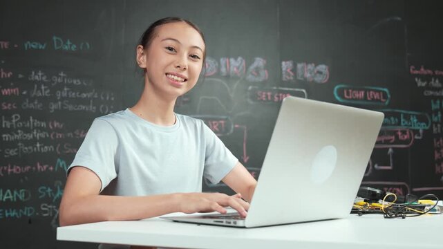 Academic high school girl working and writing code by using laptop while looking at camera at STEM technology class. Student smile to camera with electronic equipment placed on table. Edification