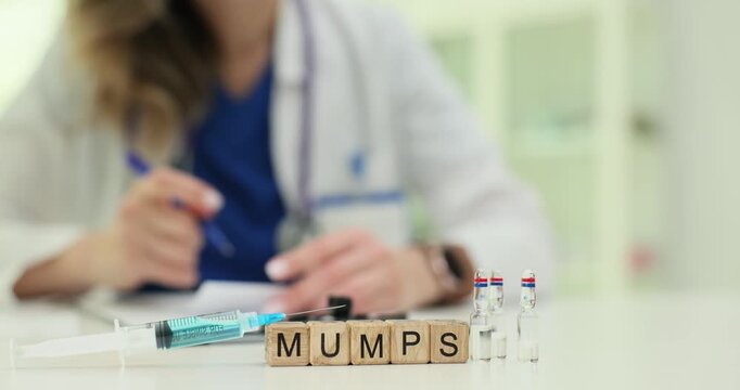 Wooden blocks spell word Mumps near vaccine vials and syringe pack. Doctor fills medical chart scheduling immunization while checking dosage labels