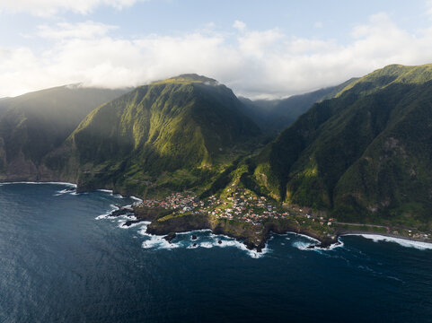 Aerial view of the coastal village of Seixal nestled between lush green mountains and the blue Atlantic Ocean under a cloudy sky in Seixal, Portugal.