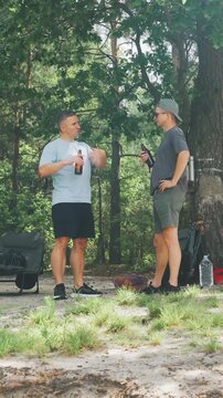 Two friends enjoy refreshing beverages outside. Vertical video. Casual gathering of two men beside flowing river in sunshine. Outdoor meetup where two men converse and share beverages by water