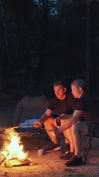 Serene evening around campfire. Calm nighttime setting with two men enjoying peaceful stargazing. Vertical video. Quiet outdoor environment with individuals relaxing by campfire beneath pine trees