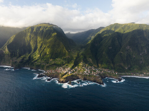 Aerial view of the coastal village of Seixal nestled between lush green mountains and the Atlantic Ocean with white waves crashing against the rocky shore Seixal, Portugal.