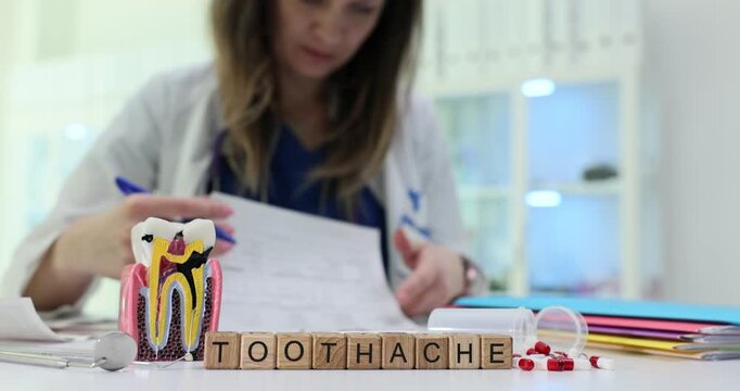 Wooden blocks spell word Toothache near tooth model and tablets on desk. Doctor reads chart recommending pain relief while scheduling dental check