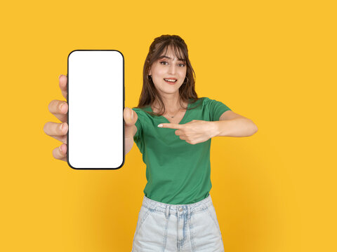 Smiling young woman holding smartphone toward camera and pointing at blank screen mockup, promoting mobile app or service. Isolated on bright yellow background with copy space for advertising.