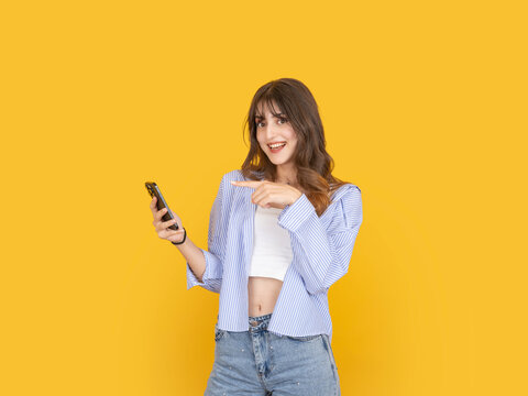Happy young woman holding smartphone and pointing at screen with cheerful expression, promoting mobile content or app concept. Isolated on bright yellow background with copy space for advertising use.