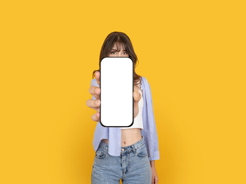 Caucasian woman holding smartphone toward camera with blank screen mock up, covering face partially, presenting mobile app, wearing striped shirt, isolated on yellow background with copy space.