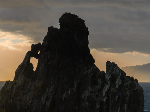 Aerial view of Ribeira da Janela sea stacks silhouetted against a dramatic sunset sky with golden light filtering through clouds over the Atlantic Ocean in Ribeira da Janela, Madeira, Portugal.