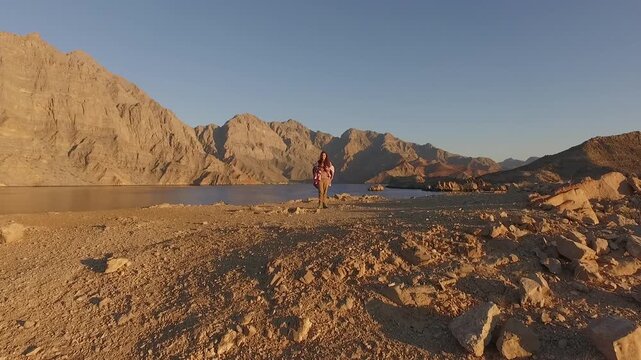Drone flying toward woman standing on rocky shore overlooking Musandam fjords near the Strait of Hormuz in Oman with dramatic desert mountains and calm sea at sunset.