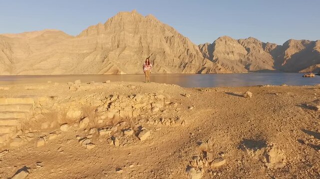 Drone flying over historic telegraph station ruins on Telegraph Island in the Strait of Hormuz Oman passing a woman explorer and revealing dramatic Musandam mountains and calm sea.