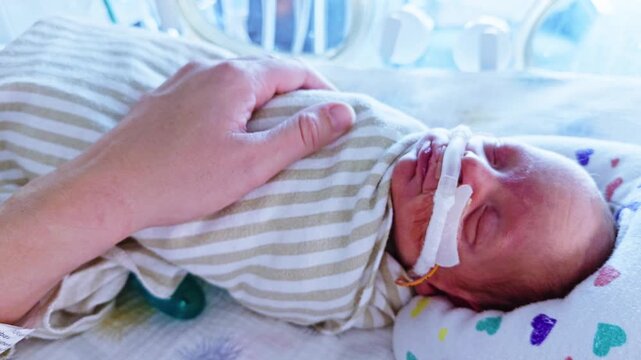 Close up of premature newborn wearing CPAP nasal prongs in NICU bassinet with parent's hand resting gently on chest.