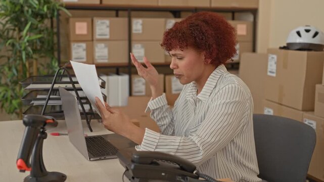 Woman reads invoice, hands holding document and raising palm in a building full of packing boxes and a laptop; frustration small business.