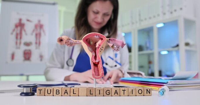 Wooden blocks spell phrase Tubal Ligation by uterus model on desk. Doctor reviews consent form explaining sterilization steps for treatment procedure