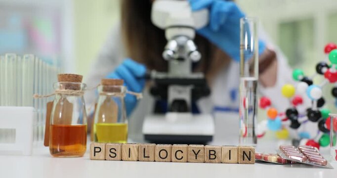 Wooden blocks spell word Psilocybin near microscope and lab samples on table. Scientist studies extract recording dosage data for controlled research