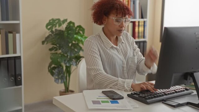 Woman typing on desktop computer with hands on keyboard, smiling and wearing glasses, red curly hair visible at desk in office building surrounded by plant and books; professional productivity.