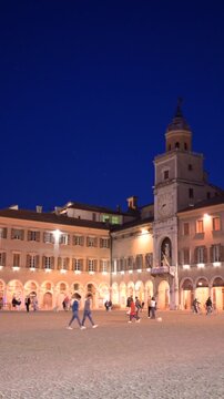 Modena, italy, april 11, 2026. Modena's piazza grande, a unesco world heritage site, glowing under night illumination. People strolling near the romanesque cathedral and ghirlandina tower