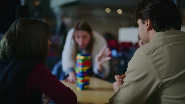 A group of young students is engaged in playing a block-stacking game at a table. They are concentrating on carefully removing and stacking colorful blocks, enjoying their leisure time.