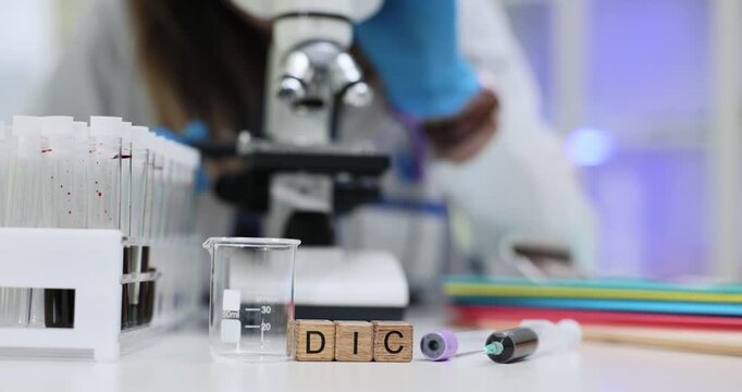 Abbreviation DIC on wooden cubes near syringe and blood samples in laboratory. Female lab assistant looks in microscope at specimen in hospital