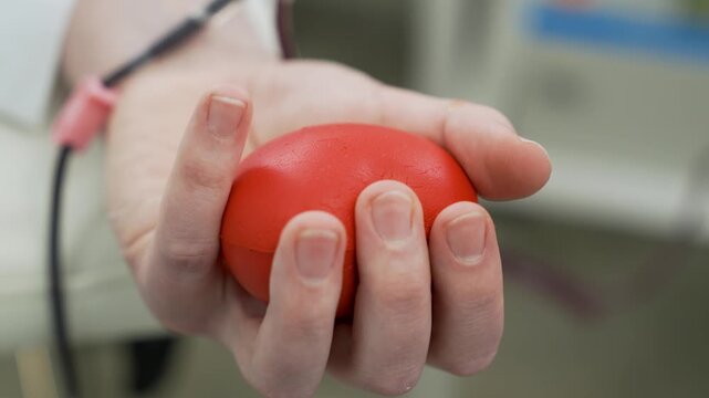 Close-up of a Blood Donor Squeezing a Red Stress Ball During Phlebotomy, Patient Hand Pumping Ball to Facilitate Blood Flow in a Medical Clinic or Hospital.