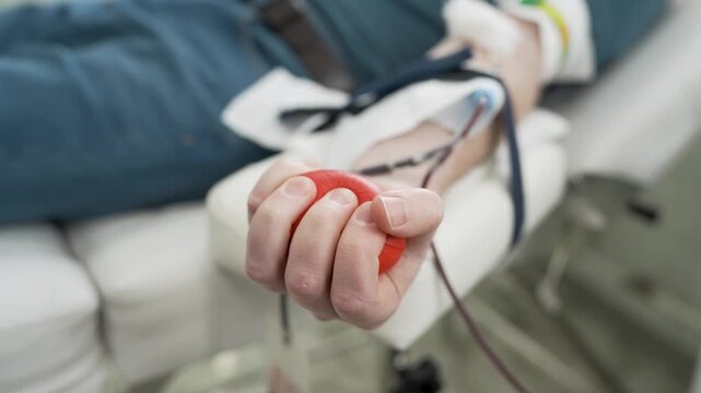 Close-up of a Blood Donor Squeezing a Red Stress Ball During Phlebotomy, Patient Hand Pumping Ball to Facilitate Blood Flow in a Medical Clinic or Hospital.