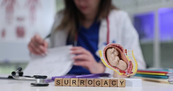 Word Surrogacy on wooden cubes near stethoscope and human fetus model on desk. Female gynecologist fills papers for parents and sur-mother in clinic