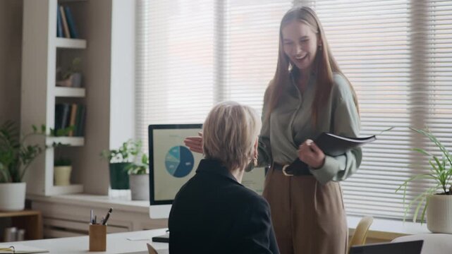 Medium shot of young Caucasian female manager entering office, greeting Black man, taking seat at desk, opening file folder and chatting with senior female colleague