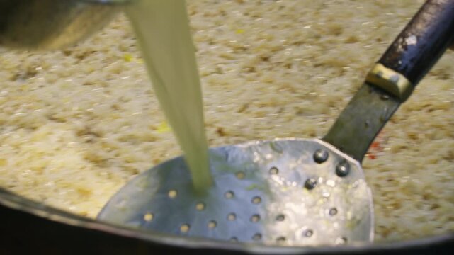 Chef pouring broth from large metal ladle over Uzbek plov in huge ancient kazan