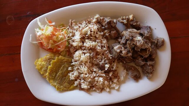 Overhead close-up shot of a typical La Guajira meal featuring stewed goat, fluffy rice, crispy patacones, and fresh salad, beautifully plated on a rustic wooden table in natural daylight.