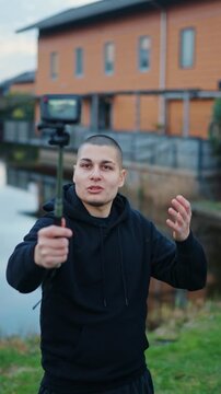 Young male influencer vlogging with an action camera on a selfie stick outdoors, talking to his followers on a live stream