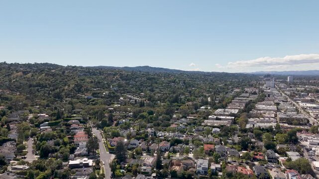 Wide aerial view over Sherman Oaks, Los Angeles showing suburban neighborhoods, tree lined streets and distant hills under bright sunlight. Calm residential cityscape in Southern California.