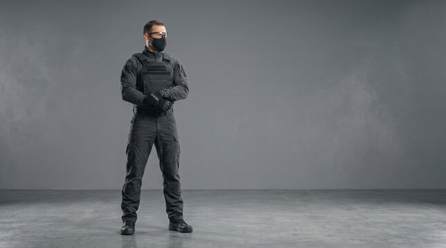 A security guard in black tactical uniform and face mask stands with arms crossed on a grey background