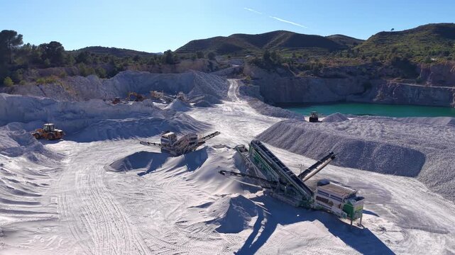 Gypsum open-pit quarry with mobile screener machines and conveyor belts processing white mineral, wheel loaders operating among large stockpiles, turquoise water pool and rocky hills in background