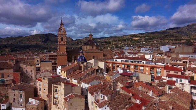Elevated view of Chelva, Spain showing terracotta rooftops, a baroque church tower and blue-tiled dome, surrounded by green hills under a cloudy sky