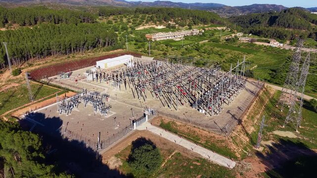Overhead view of a large electrical substation surrounded by green forest and farmland, featuring transformers, transmission towers, and complex wiring infrastructure