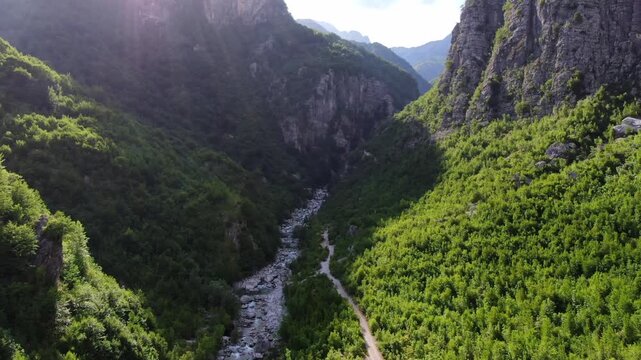 Dramatic aerial view flying slowly through a deep green valley in Theth National Park, Albania, with a river and path below