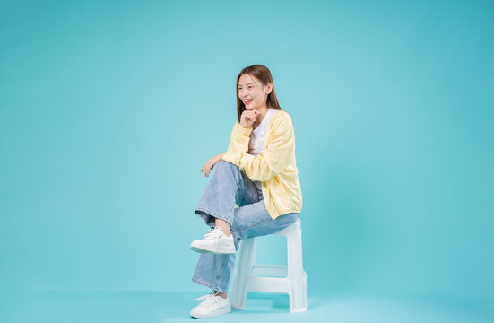 Happy Young Asian Woman Sitting and Thinking on Turquoise Background. Portrait of Smiling Female with Copy Space for Inspiration, Success, and Positive Thinking Concept
