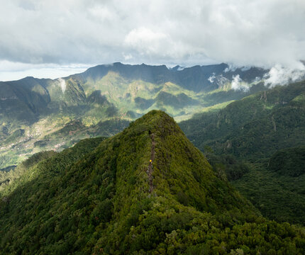 Aerial view of the PR17 Pinaculo e Folhadal hiking trail along a lush green mountain ridge with a hiker in yellow under a cloudy sky in Serra de Agua, Madeira, Portugal.
