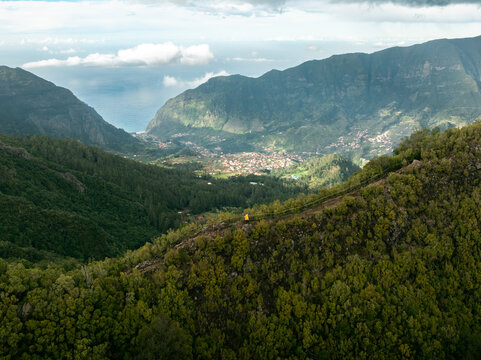 Aerial view of PR17 Pinaculo e Folhadal hiking trail with a person in a yellow jacket on a ridge overlooking the valley and ocean Serra de Agua, Madeira, Portugal.