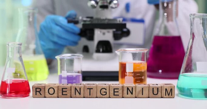 Word Roentgenium on wooden cubes near flasks with color fluids in lab. Female assistant uses microscope to analyze material specimens at desk