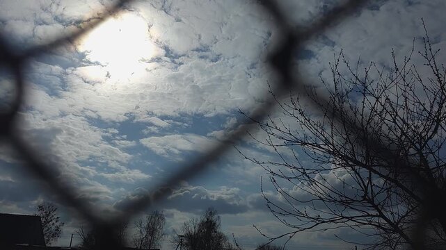 View of bright sun through a chain-link fence against blue sky with white clouds and bare tree branches on the side, concept of restriction and hope