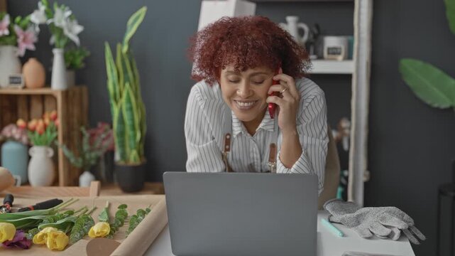 Woman florist holds smartphone to ear and leans over laptop while arranging stems on worktable in studio; small business happiness.