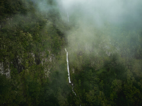 Aerial view of PR17 Pinaculo e Folhadal waterfall cascading down a steep cliff surrounded by dense green forest and thick mist Serra de Agua, Madeira, Portugal.