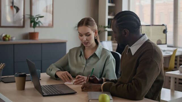 Medium shot of gen Z Caucasian and Black colleagues brainstorming at work in modern open space office, sitting at desk, looking at laptop and throwing away papers with bad ideas