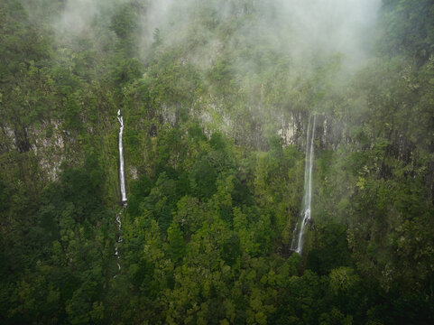 Aerial view of PR17 Pinaculo e Folhadal waterfalls cascading down steep cliffs through dense green forest and mist in Serra de Agua, Madeira, Portugal.