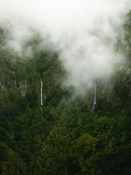 Aerial view of the PR17 Pinaculo e Folhadal area with waterfalls cascading down steep cliffs through dense green forest and thick fog in Serra de Agua, Madeira, Portugal.