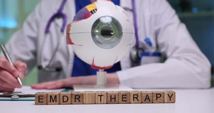 Wooden blocks spell phrase EMDR Therapy near eyeball model on desk. Therapist writes session notes planning trauma processing steps for patient care