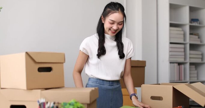 Young asian woman entrepreneur working from home, cheerfully packing merchandise into cardboard boxes for online orders, managing her small business with a laptop