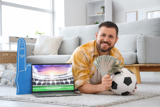 Happy young man with soccer ball and laptop lying on floor at home. Sports bet concept